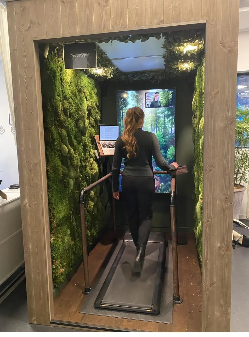 A person walking on the pad inside a Nature Station pod during a video meeting, moss walls on both sides and a forest on the screen